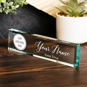 Puede incluir: Placa de escritorio rectangular de vidrio con el texto "Your Name" y "Your Title" en fuente cursiva. Un área de logotipo circular dice "Your Logo Goes Here". La placa de identificación está sobre una superficie de madera.