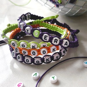 May include: Four colorful friendship bracelets with the words "Spooky" and "Era" spelled out in white beads. The bracelets are made with purple, green, orange, and white string.