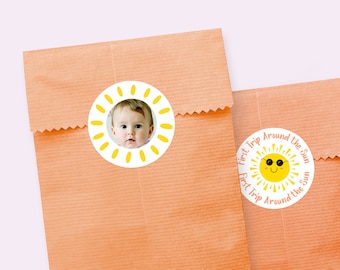 May include: A white round sticker with a yellow sun and the text "First Trip Around the Sun" in orange. The sun has sunglasses and a smiling face.