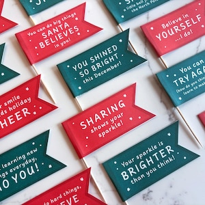 May include: A collection of red and teal flag-shaped paper decorations with white text and star accents. The flags feature positive messages like "CHEER," "BELIEVE," and "SHARING." Each flag is attached to a wooden stick.
