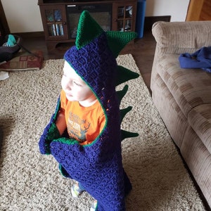 Puede incluir: Un ni&ntilde;o lleva una manta de capucha de drag&oacute;n de crochet azul y verde. La manta tiene una cola larga y p&uacute;as. El ni&ntilde;o lleva una camisa naranja con un gr&aacute;fico de ciervo y el texto "New England 1887 Camping".