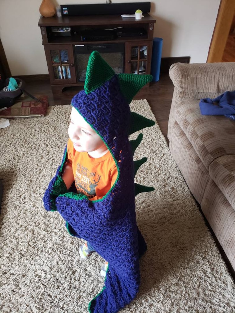 Puede incluir: Un ni&ntilde;o lleva una manta de capucha de drag&oacute;n de crochet azul y verde. La manta tiene una cola larga y p&uacute;as. El ni&ntilde;o lleva una camisa naranja con un gr&aacute;fico de ciervo y el texto "New England 1887 Camping".