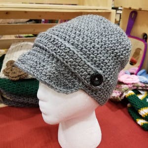 Op de afbeelding: Een grijze gehaakte beanie met een rand en een zwarte knoop. De beanie zit op een wit mannequin hoofd.