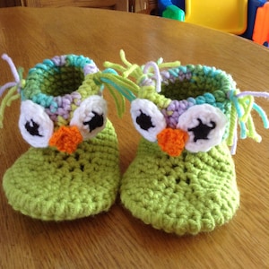 May include: A pair of green crocheted owl baby booties with white, black, and orange felt accents. The booties have a fringe of yarn around the top.