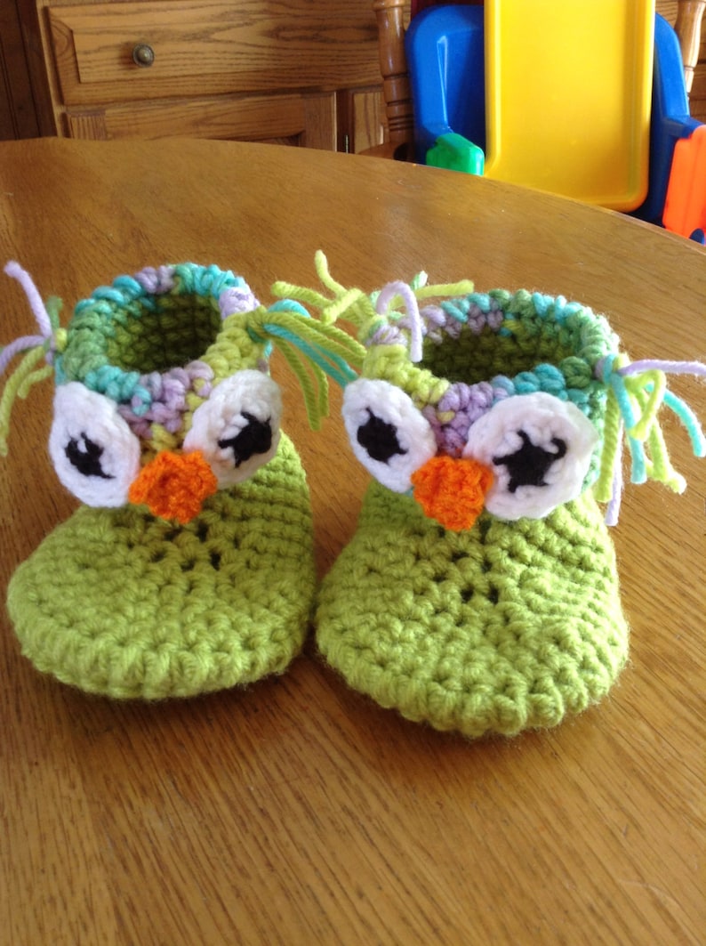 May include: A pair of green crocheted owl baby booties with white, black, and orange felt accents. The booties have a fringe of yarn around the top.
