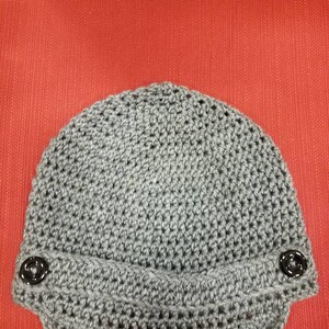 Op de afbeelding: Grijze gebreide beanie met een rand en twee zwarte knopen aan de zijkanten.
