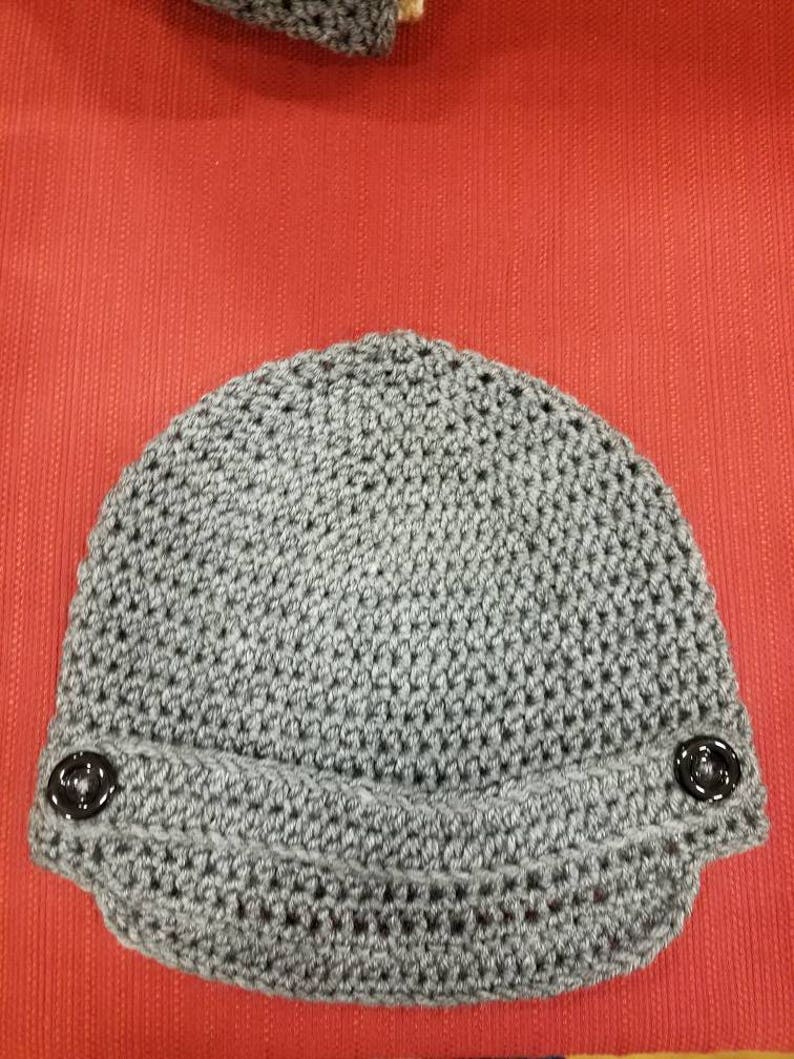 Op de afbeelding: Grijze gebreide beanie met een rand en twee zwarte knopen aan de zijkanten.