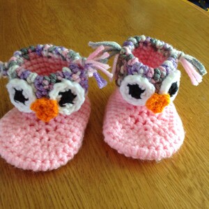 May include: A pair of pink crocheted baby booties with owl faces. The booties have white eyes, black pupils, and orange beaks. The booties have a gray, pink, and purple crocheted trim around the top.