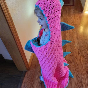 Puede incluir: Una manta de dinosaurio de crochet rosa y azul con capucha. La manta est&aacute; hecha con un punto texturizado y tiene una cola con p&uacute;as.