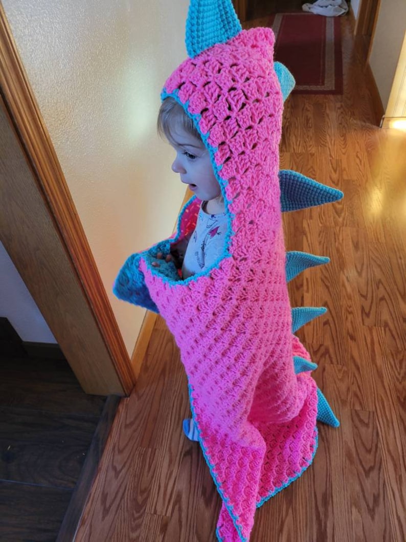 Puede incluir: Una manta de dinosaurio de crochet rosa y azul con capucha. La manta est&aacute; hecha con un punto texturizado y tiene una cola con p&uacute;as.