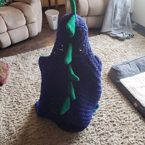 Puede incluir: Una manta de dinosaurio de crochet azul y verde con una cola larga y p&uacute;as. La manta est&aacute; doblada y sentada sobre una alfombra marr&oacute;n claro.