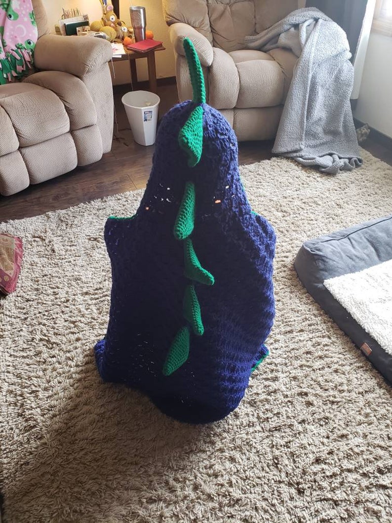 Puede incluir: Una manta de dinosaurio de crochet azul y verde con una cola larga y p&uacute;as. La manta est&aacute; doblada y sentada sobre una alfombra marr&oacute;n claro.