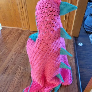 Puede incluir: Una manta de dinosaurio de crochet rosa y turquesa con capucha. La manta est&aacute; hecha con un punto texturizado y tiene una cola larga y p&uacute;as.
