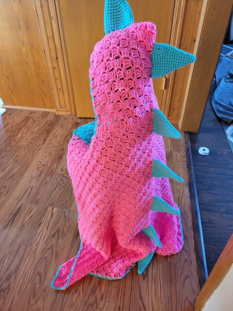 Puede incluir: Una manta de dinosaurio de crochet rosa y turquesa con capucha. La manta est&aacute; hecha con un punto texturizado y tiene una cola larga y p&uacute;as.