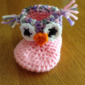 May include: Pink crocheted baby booties with an owl design. The owl has white eyes with black pupils, an orange beak, and a purple, pink, and gray crocheted crown.