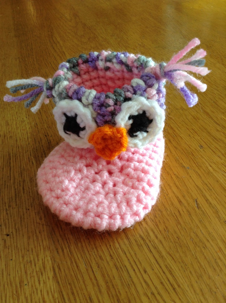 May include: Pink crocheted baby booties with an owl design. The owl has white eyes with black pupils, an orange beak, and a purple, pink, and gray crocheted crown.