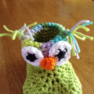 May include: Green crocheted owl baby booties with white eyes, black pupils, and an orange nose. The booties have colorful yarn accents.