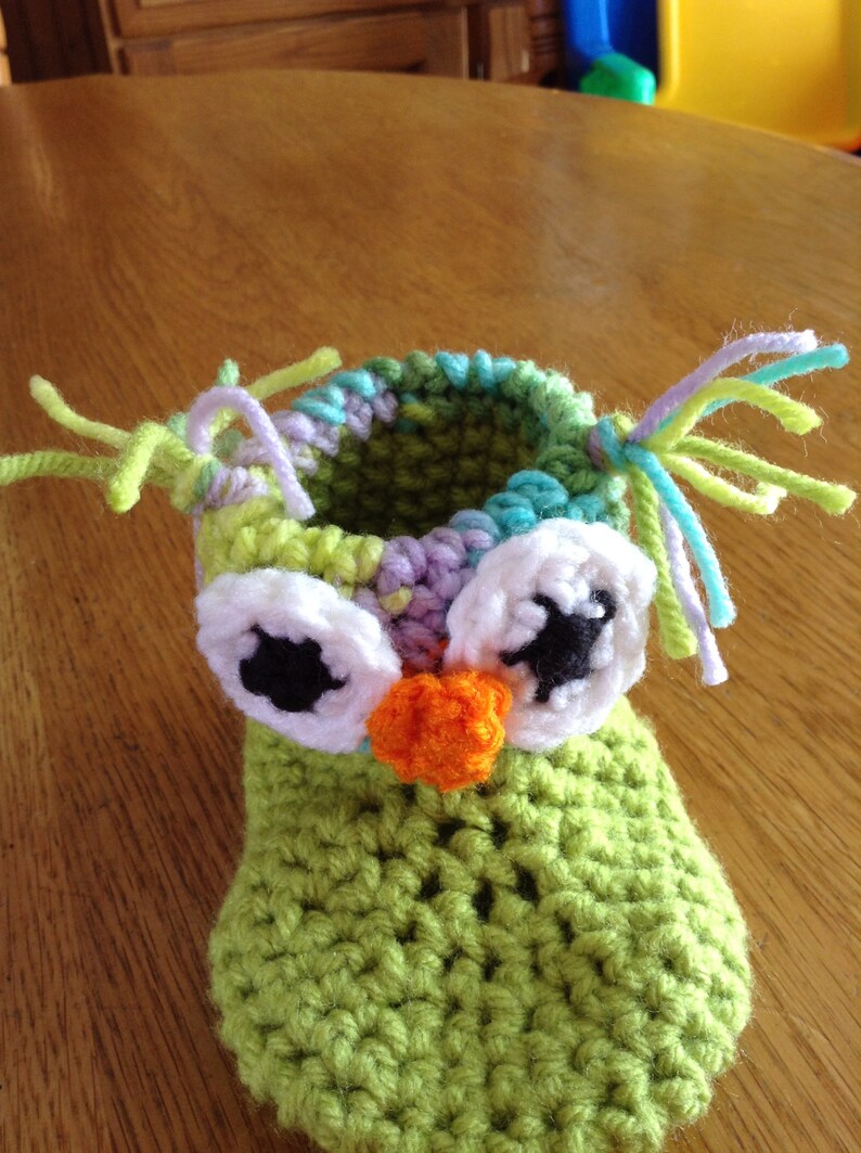 May include: Green crocheted owl baby booties with white eyes, black pupils, and an orange nose. The booties have colorful yarn accents.