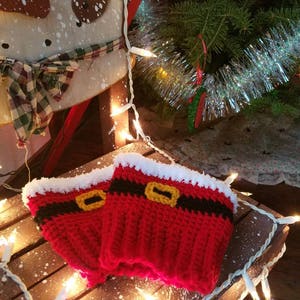 May include: A pair of red crocheted Santa boot cuffs with a black belt and a gold buckle. The cuffs have a white fuzzy trim.