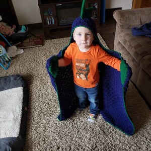 Puede incluir: Un ni&ntilde;o peque&ntilde;o lleva una manta con capucha de crochet azul y verde con una cabeza de dinosaurio en la parte superior. El ni&ntilde;o lleva una camiseta naranja con el texto "Always on the Trail Since 1889 Carhartt Hunt the Wild" y pantalones vaqueros azules. El ni&ntilde;o est&aacute; de pie sobre una alfombra beige.