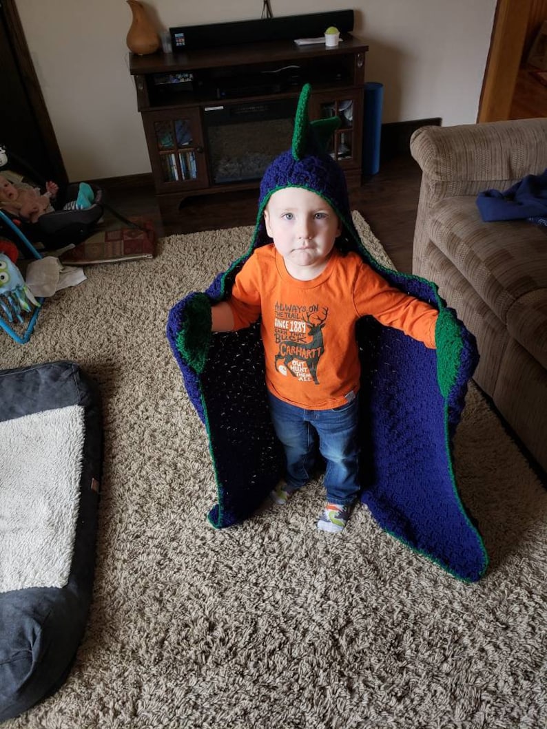 Puede incluir: Un ni&ntilde;o peque&ntilde;o lleva una manta con capucha de crochet azul y verde con una cabeza de dinosaurio en la parte superior. El ni&ntilde;o lleva una camiseta naranja con el texto "Always on the Trail Since 1889 Carhartt Hunt the Wild" y pantalones vaqueros azules. El ni&ntilde;o est&aacute; de pie sobre una alfombra beige.