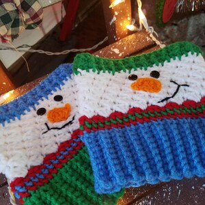 May include: Two crocheted snowman mug cozies with white, blue, green, and red yarn. The cozies have black button eyes, orange yarn noses, and a stitched smile.