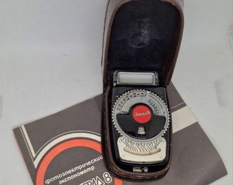May include: Vintage Leningrad 8 light meter in a brown leather case, with a black and silver dial and a red center. The meter is open, revealing the dial. A gray brochure with Russian text is below.