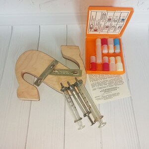 Op de afbeelding: Een vintage medische set met drie glazen spuiten en een klein oranje doosje met verschillende flesjes. De set ligt op een licht houten oppervlak.