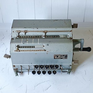 Op de afbeelding: Een vintage, grijze metalen rekenmachine met een kruk en een display dat de nummers 999969331100 0000000 laat zien. De machine heeft een reeks genummerde toetsen langs de onderkant.
