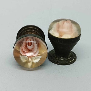 May include: Two vintage cabinet knobs featuring a rose design. The knobs are made of metal and have a clear glass top with a pink rose design. The knobs are in good condition and would be a great addition to any home.