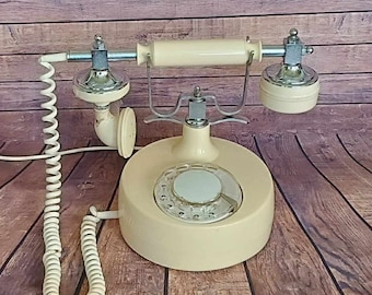 Pode incluir: Um telefone de disco vintage com corpo de cor creme e detalhes prateados. O telefone tem um cabo espiralado e um disco rotativo com números pretos.