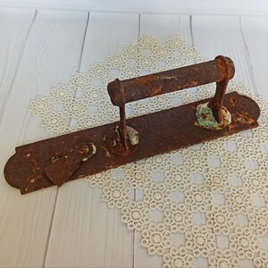 May include: A rusty metal door handle with a long, rectangular base and a curved handle. The handle is attached to the base with screws and has a decorative design. The handle is covered in rust and has a few spots of green paint.