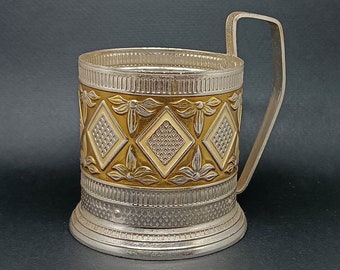 May include: A silver and gold metal cup holder with a geometric pattern. The cup holder has a handle and a wide base.