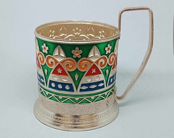 May include: Silver-plated cup holder with a green, orange, blue, and white geometric design. The holder has a handle and a decorative base.