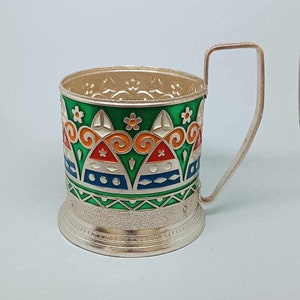 May include: Silver-plated cup holder with a green, orange, blue, and white geometric design. The holder has a handle and a decorative base.