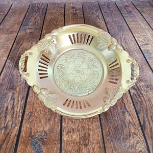 Puede incluir: Una bandeja dorada decorativa con un diseño floral y de hojas. La bandeja tiene un centro redondo con un patrón floral y una serie de cortes verticales alrededor del borde. Cuenta con asas y detalles decorativos de hojas y bayas.