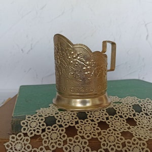 May include: A gold-colored metal cup holder with an ornate floral design. The holder is sitting on a stack of books covered in a lace doily.
