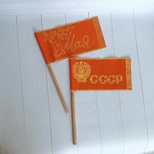 Könnte beinhalten: Zwei orangefarbene Stofffahnen auf Holzstöcken. Die obere Fahne trägt den Text "1. Mai" in goldenen Buchstaben mit einem floralen Design. Die untere Fahne trägt den Text "CCCP" in goldenen Buchstaben mit einem goldenen Emblem.