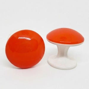 May include: Two mushroom-shaped knobs with smooth, glossy orange caps and solid white bases. One knob is positioned to show the top of the cap, while the other is turned to the side to reveal the white stem and the underside of the orange cap.