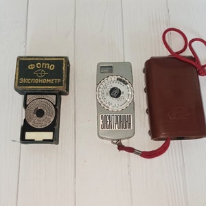 Puede incluir: Tres exposímetros vintage, uno en una caja negra con el texto "Фото Экспонометр", otro en una caja plateada con el texto "Электроника" y otro en una funda de cuero marrón con una correa roja.