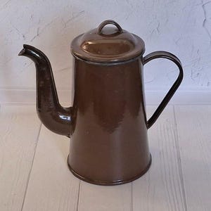 以下が含まれることがあります： 蓋と取っ手がついた茶色のエナメル製のコーヒーポット。ポットは白い木製の表面の上に置かれています。
