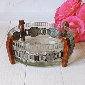 May include: A clear glass bowl with a decorative silver trim and wooden accents. The bowl is sitting on a white surface.