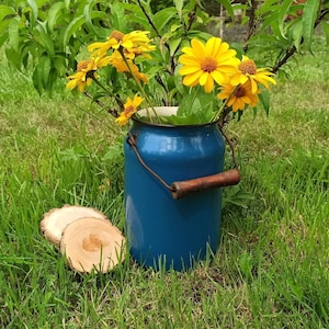 Puede incluir: Una jarra de esmalte azul con un asa de madera llena de flores amarillas. La jarra está sentada en un césped verde.
