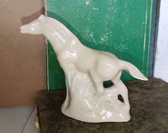 May include: A white porcelain figurine of a horse standing on a rocky base. The horse is in a running pose with its head turned to the side.