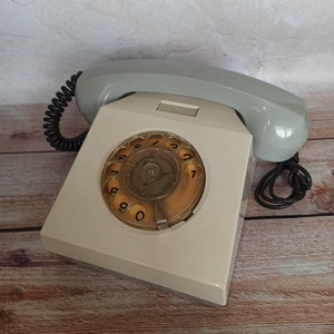 May include: A white rotary dial phone with a gray handset and a black coiled cord. The phone has a circular dial with black numbers 0-9.