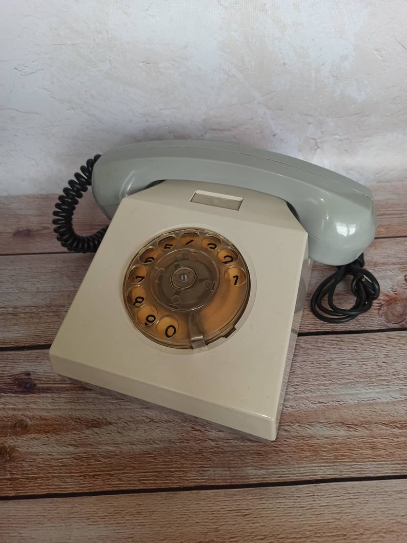 May include: A white rotary dial phone with a gray handset and a black coiled cord. The phone has a circular dial with black numbers 0-9.
