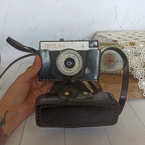 Puede incluir: Una cámara vintage negra con una funda de cuero. La cámara tiene una lente plateada con una etiqueta roja "SMENA 8M" en la parte delantera.
