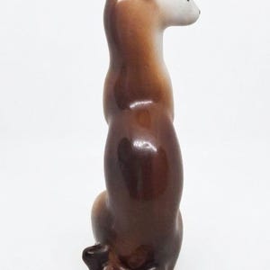 K&ouml;nnte beinhalten: Eine gl&auml;nzende Keramikfigur eines schlanken, langgestreckten Tieres, wahrscheinlich ein Frettchen oder Wiesel, in sitzender Position. Die Figur hat eine glatte Farbverlaufsoberfl&auml;che, die von einem dunklen Braun am K&ouml;rper zu einem hellen Kopf &uuml;bergeht.