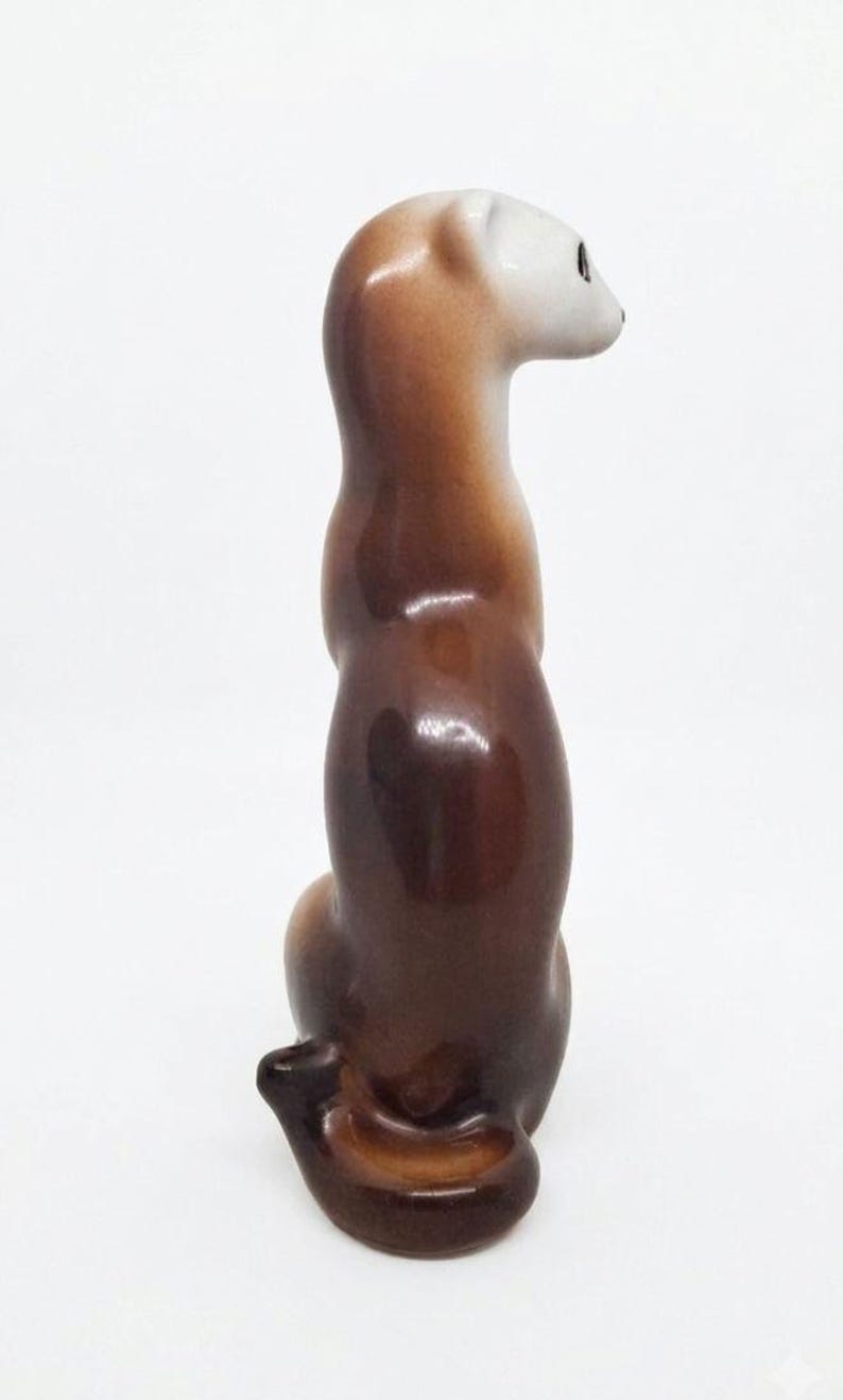 K&ouml;nnte beinhalten: Eine gl&auml;nzende Keramikfigur eines schlanken, langgestreckten Tieres, wahrscheinlich ein Frettchen oder Wiesel, in sitzender Position. Die Figur hat eine glatte Farbverlaufsoberfl&auml;che, die von einem dunklen Braun am K&ouml;rper zu einem hellen Kopf &uuml;bergeht.
