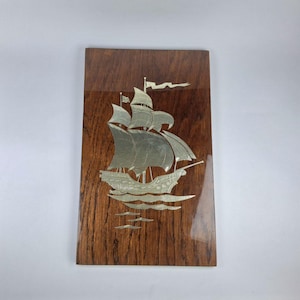 May include: A rectangular wooden plaque with a silver-colored ship design. The ship has multiple sails and is depicted on water. The wood has a dark brown grain pattern and a glossy finish, creating a vintage nautical theme.